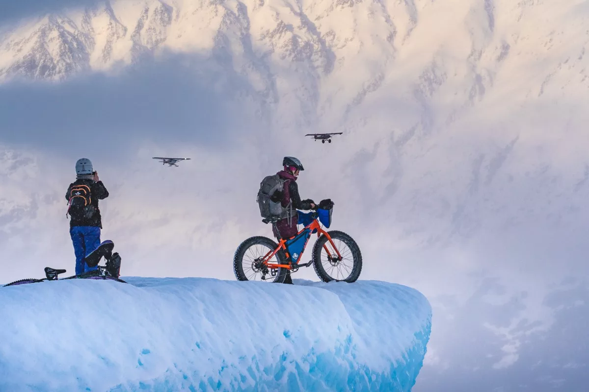 bikerumor pic of the day two fat bike cyclists on the edge of an icy overhang looking out over a glacier with two small aircraft in the distance.