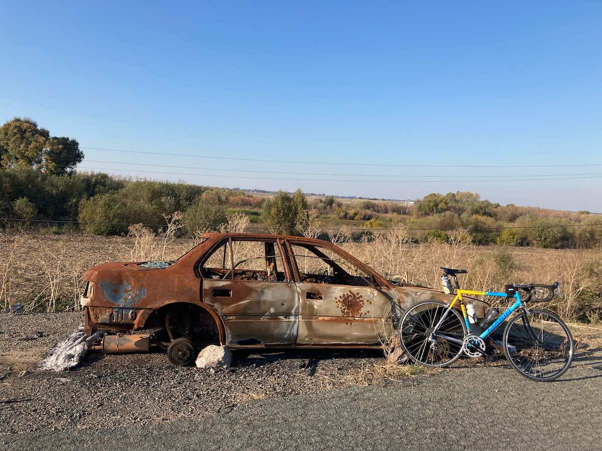 bikerumor pic of the day a bicycle leans against a rusty shell of a car on a gravel road next to a field of golden grasses