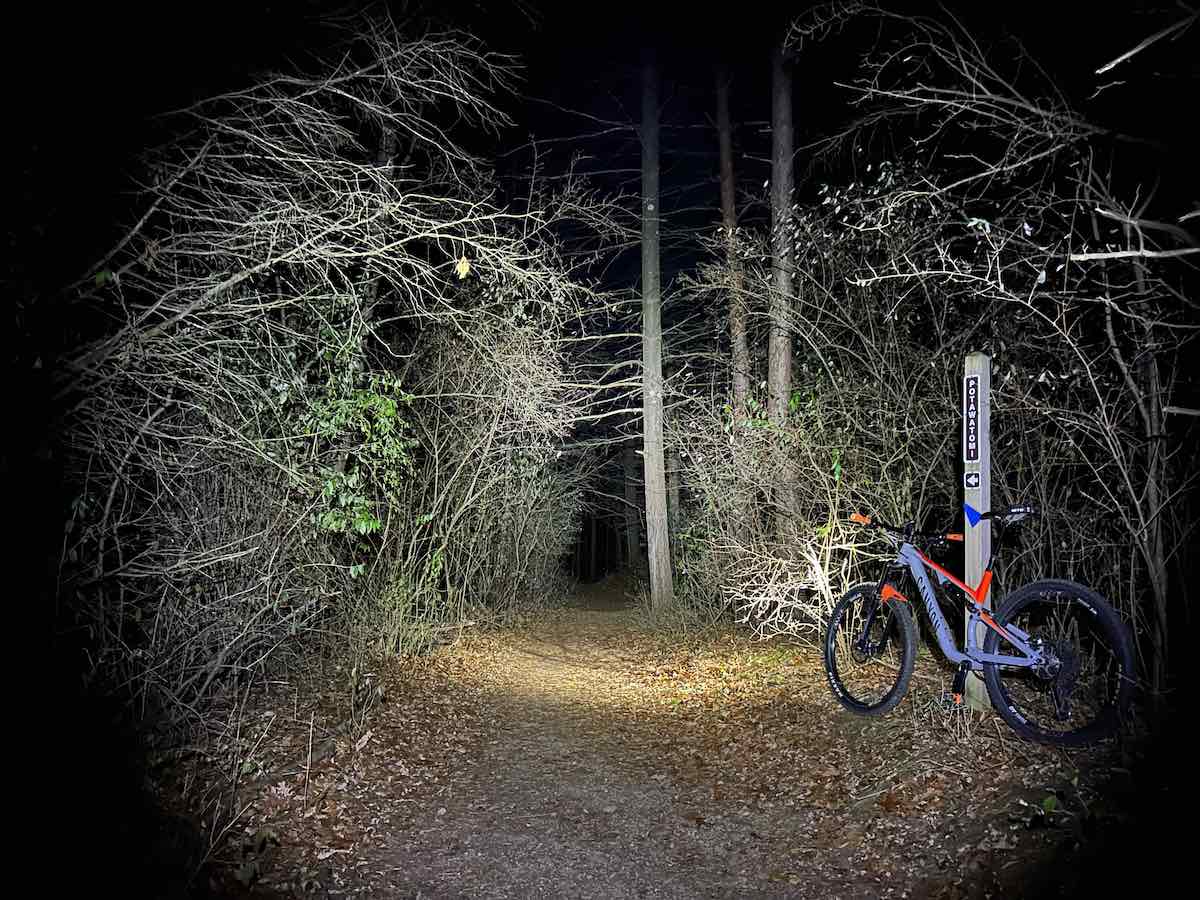 bikerumor pic of the day a mountain bike with lights on is leaning against a wood post on a trail at night the trees are bare and there are leaves on the ground.