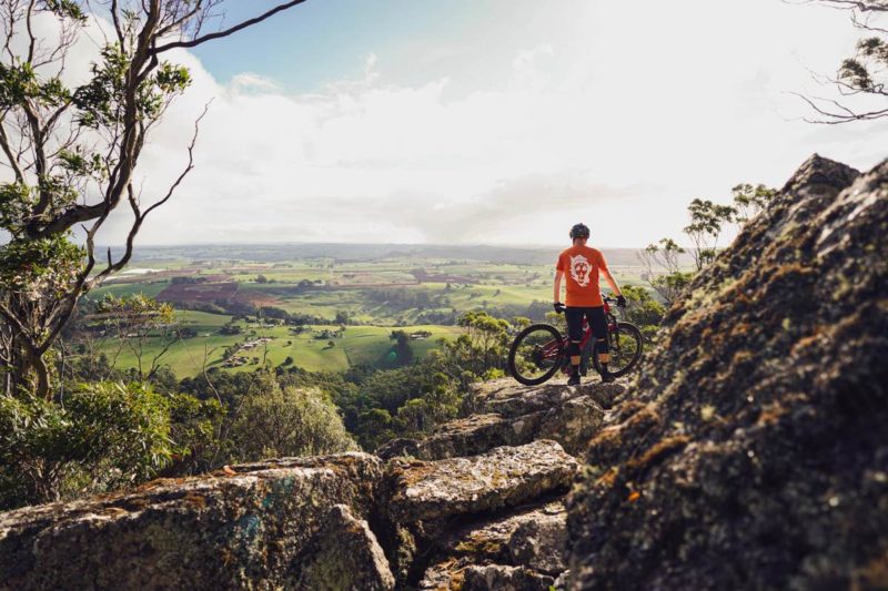 a mountain biker standing with their bike at the top of a rocky outcropping overlooking a vast green valley