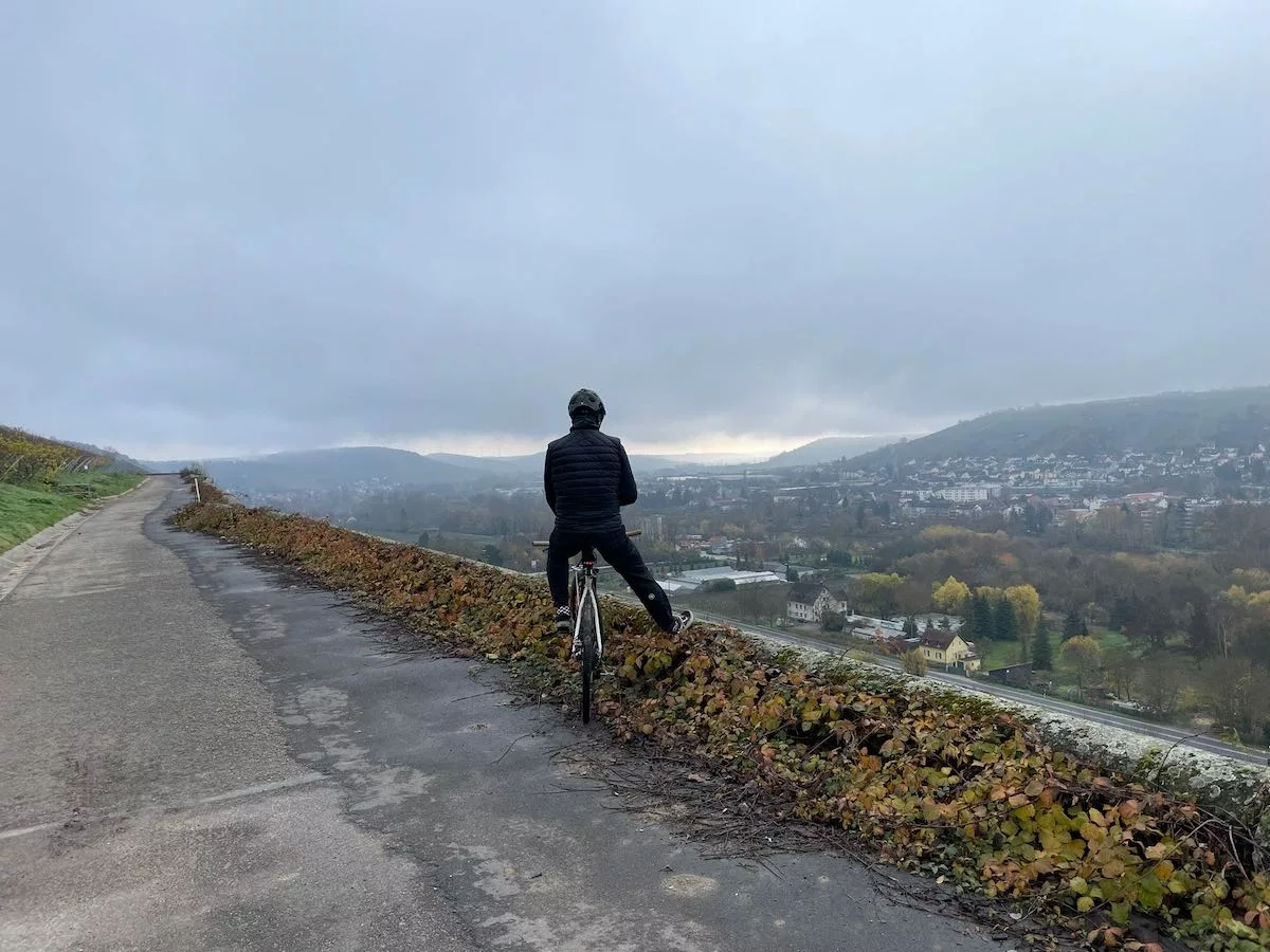 bikerumor pic of the day a cyclist pauses along a road overlooking a valley, the sky is cloudy and the sun is peeing out along the horizon.
