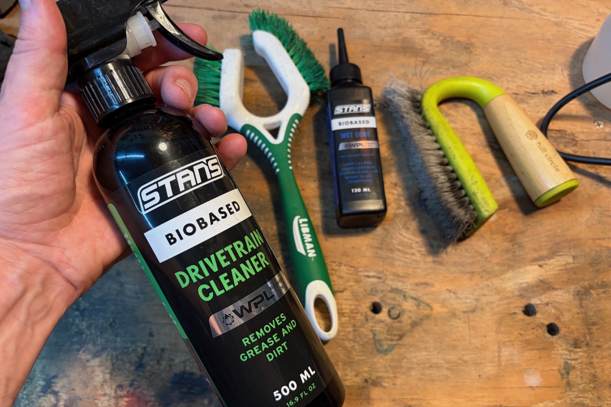 holding the bottle of Stan's Biobased Drivetrain Cleaner with brushes on a workbench behind.