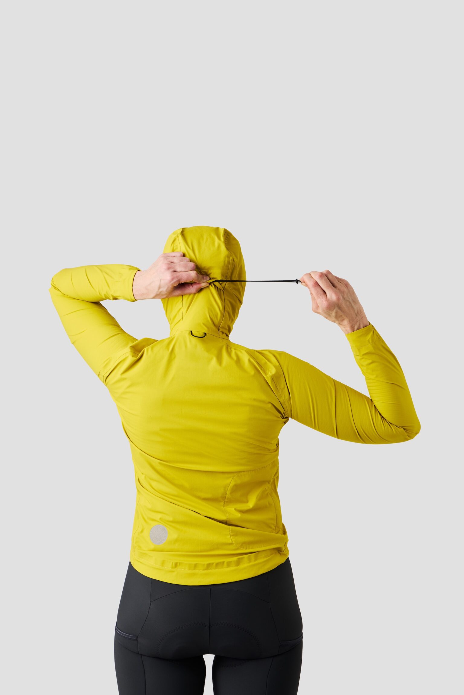 studio image of a woman wearing the Ornot Fog eater Hooded Rain Jacket tightening the hood