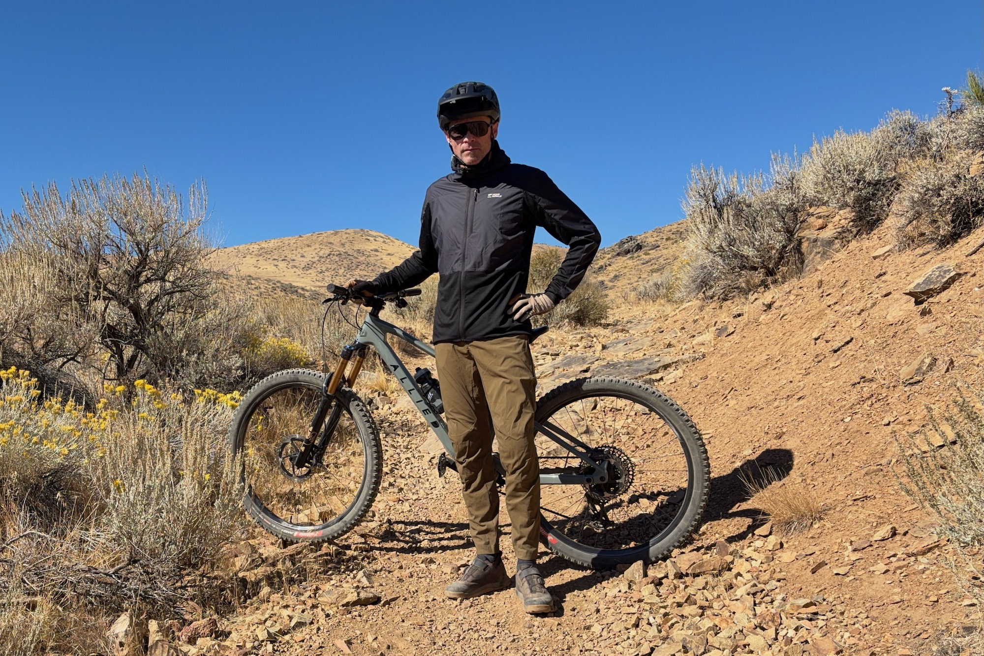 standing with a mountain bike on a trail while wearing the mons Royale Diversion pants and wind jacket.